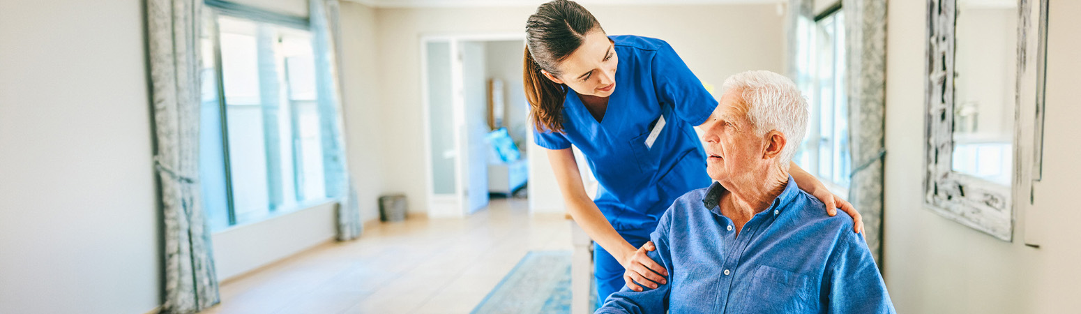 Home health nurse assisting an elderly male patient in a bright, comfortable home setting.