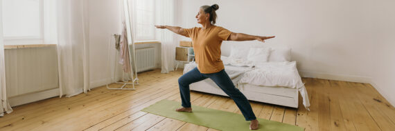 Older woman practicing yoga in a Warrior II pose at home, demonstrating integrative medicine's use of yoga for holistic health.