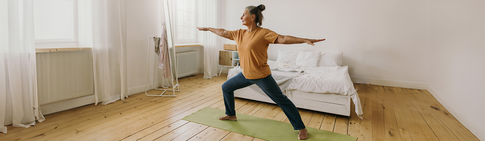 Older woman practicing yoga in a Warrior II pose at home, demonstrating integrative medicine's use of yoga for holistic health.