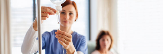 Nurse adjusting IV drip with a patient in the background, illustrating the choice between midline vs PICC lines in care.