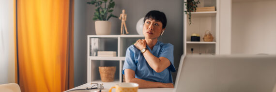 Healthcare professional in scrubs sitting at a desk, holding their shoulder and appearing reflective.