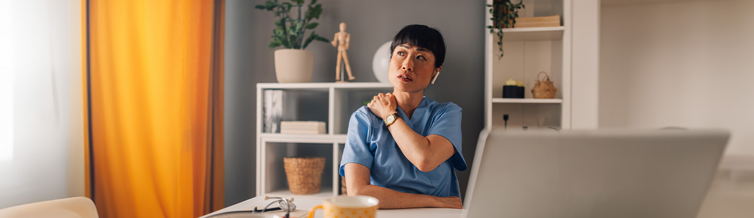 Healthcare professional in scrubs sitting at a desk, holding their shoulder and appearing reflective.