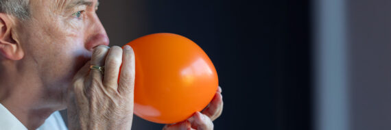 A man inflating an orange balloon, demonstrating a voice therapy exercise to improve breath support and vocal function.