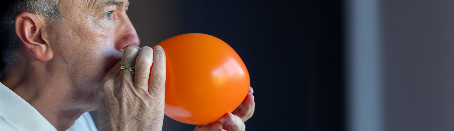 A man inflating an orange balloon, demonstrating a voice therapy exercise to improve breath support and vocal function.