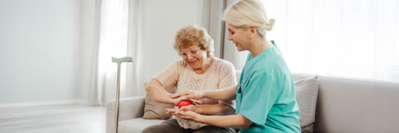 A healthcare professional helps an elderly woman with a hand exercise using a red therapy ball in a home setting.