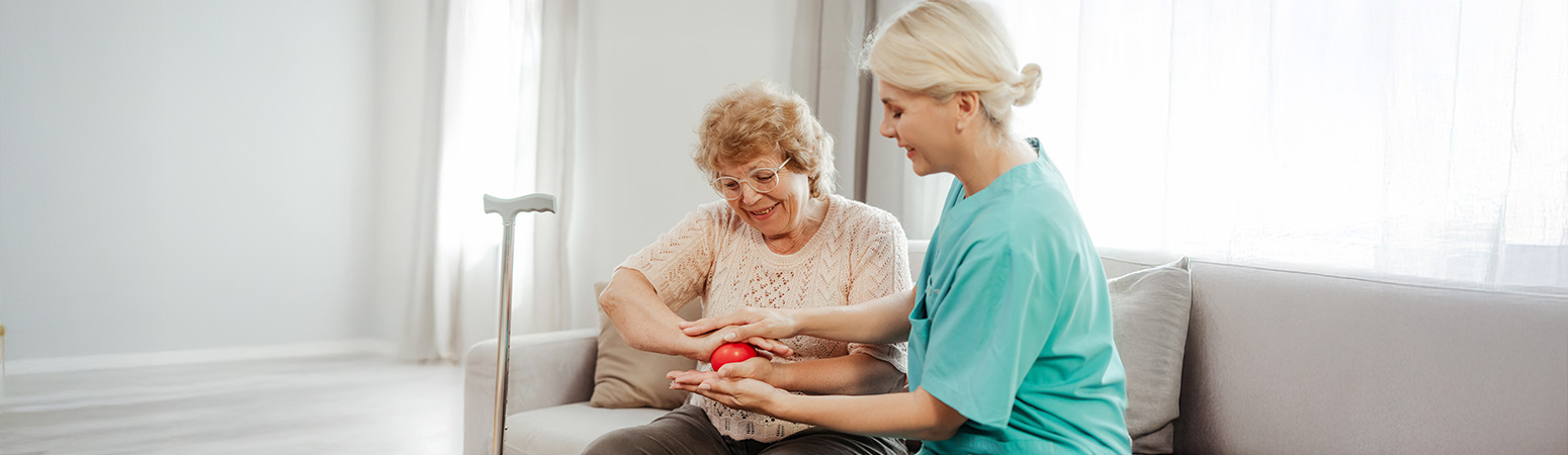 A healthcare professional helps an elderly woman with a hand exercise using a red therapy ball in a home setting.