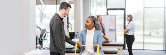 Physical therapist encouraging a seated female patient using a resistance band, with a supportive clinical setting in the background.