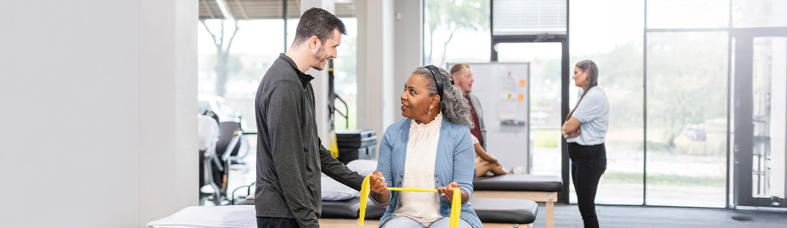 Physical therapist encouraging a seated female patient using a resistance band, with a supportive clinical setting in the background.