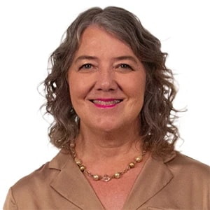 Person with curly shoulder-length hair, pearl necklace, and beige shirt against a white background.